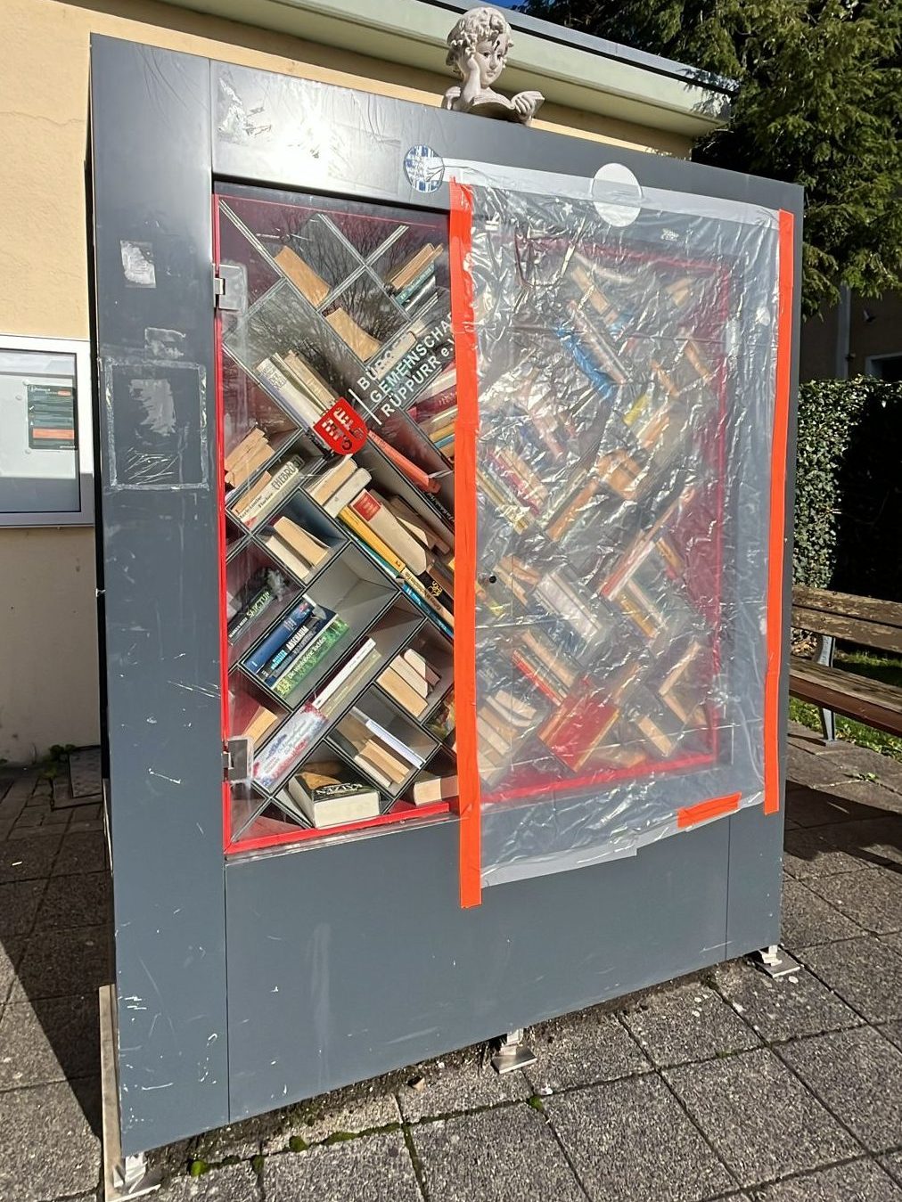 Foto des Bücherschranks. Tür auf der Vorderseite fehlt und statt dessen ist der Schrank mit einer Folie verschlossen.