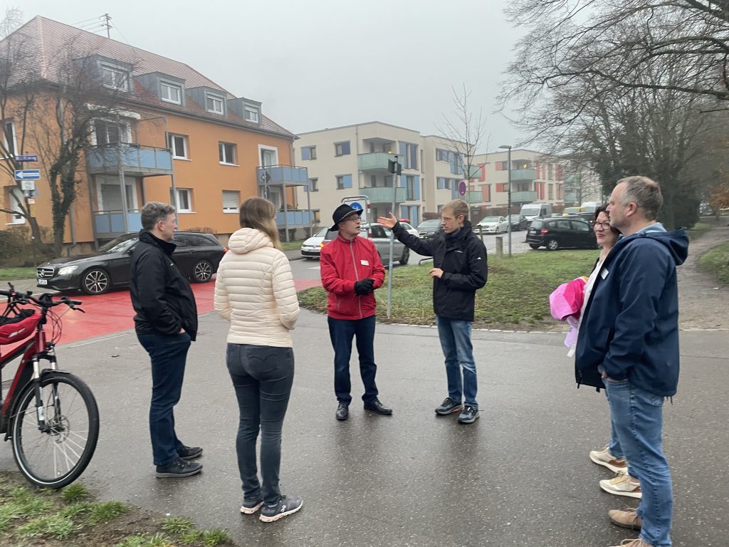 Foto vom Ortstermin. Man sieht die im Bericht genannten Person im Kreis stehen und diskutieren. Im Hintergrund sind Fahrzeuge zu sehen. Der Boden ist nass und das Foto gibt eine ungemütliche Atmosphere wieder.