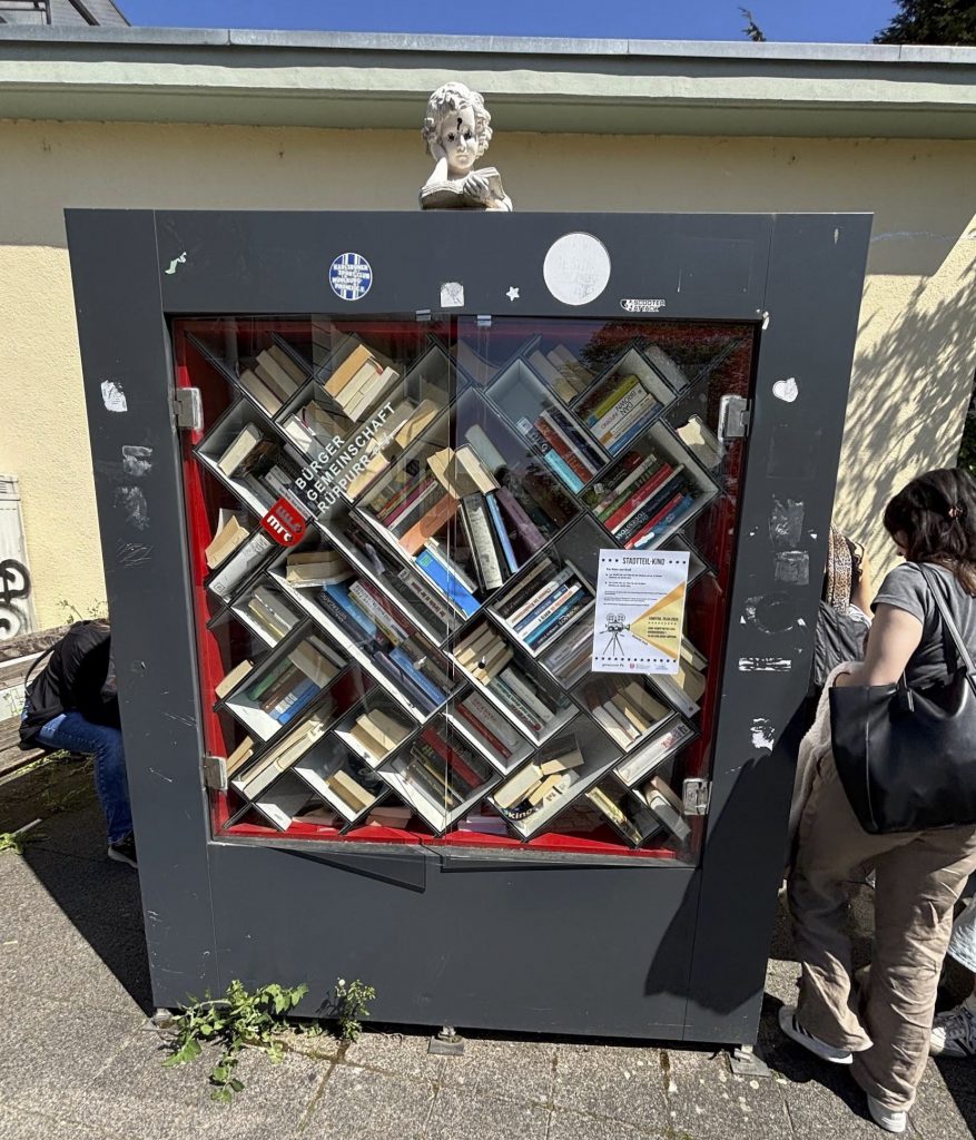 Frontansicht des Bücherschranks. Glastüren stehen leicht offen. Der Schrank sieht mitgenommen aus und ist mit zum Teil wieder entfernten Stickern beklebt. Auf dem Schrank ist die Figur eines Kindes zu sehen, welches ein Buch in den Händen hält. Die Augen wurden schwarz bemahlt.