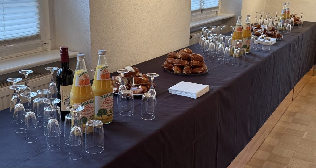 Langer Tisch vorbereitet für den Jahresempfang. Schwarze Tischdecke. Auf dem Tisch stehen Gläser, Orangensaft und Sekt. Zu Essen gibt es Laugenstangen mit Butter.