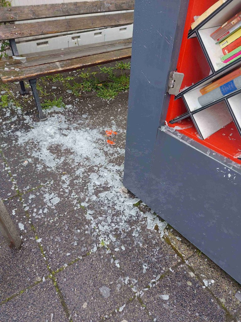 Aufnahme des linken unteren Teil des Bücherschrankes. Zerbröseldes Glas der Türscheibe liegt auf dem Boden.