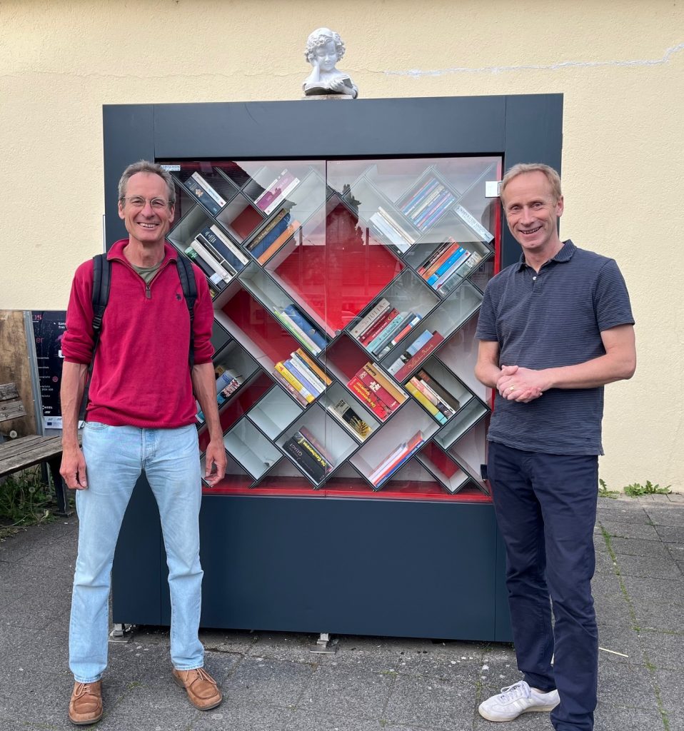 Zwei Männer stehen vor dem neu renovierten Bücherschrank. Bücher wurden bereits wieder einsortiert. Der Schrank wurde komplett neu gestrichen und neue Scheiben wurden eingesetzt. Alles sieht schön neu und sauber aus.