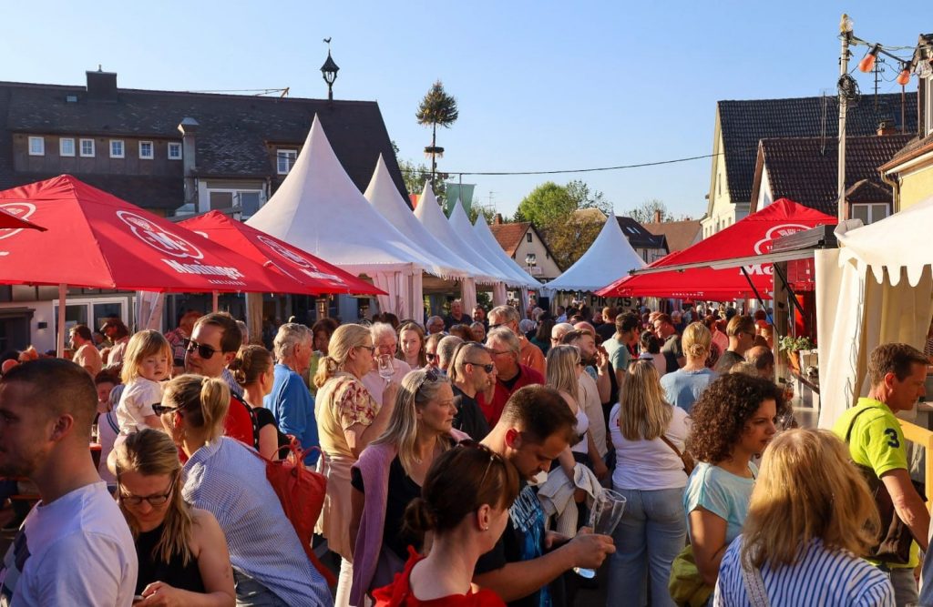 Foto vom Maibaumfest - Blick auf die Rastatter Straße, gefüllt mit Menschen die sich durch die Straße drängen. Im Hintergrund ist der Maibaum zu sehen. Davor Zelte und Schirme mit Bierbänken.
