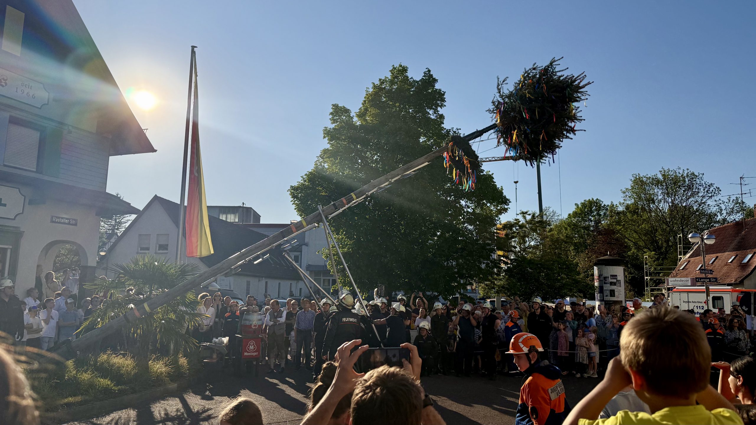 Foto vom Aufstellen des Maibaumes. Ca. 50 Zuschauer sind im Hintergrund zu sehen. Die Freiwillige Feuerwehr Rüppurr ist gerade dabei den Baum mit langen Metallstäben in die Senkrechte zu stellen. Auf dem Bild ist der Baum nur zur Hälfte errichtet.