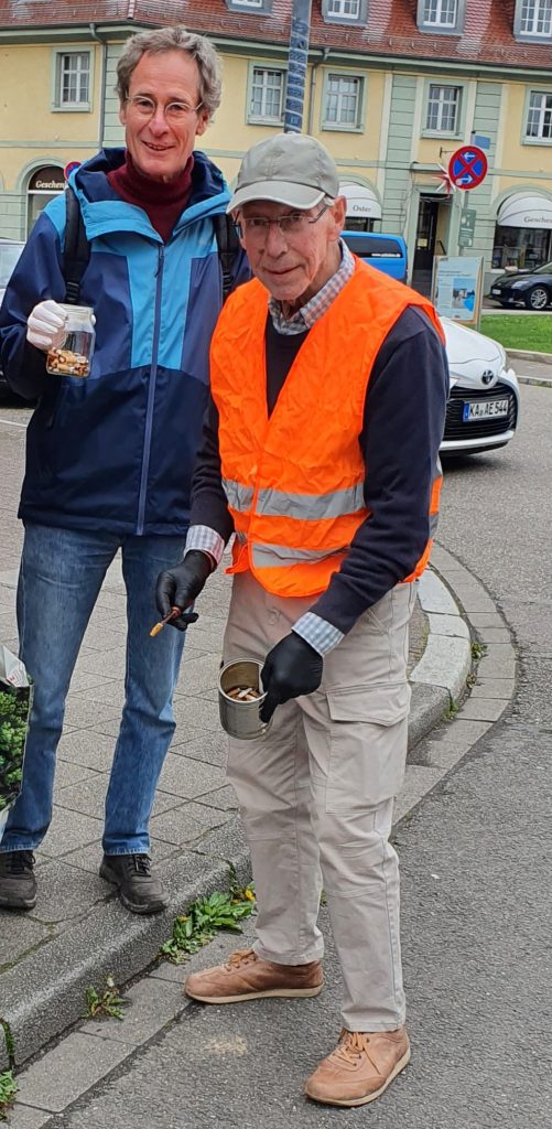 Foto von 2 Männern die am Ostendorfplatz stehen und dabei sind Zigarettenkippen zu sammeln. Der Mann auf der rechten Seite hat eine orangene Sicherheitsweste an. In den Händen halten sie Gefäße mit bereits aufgesammelten Kippen.