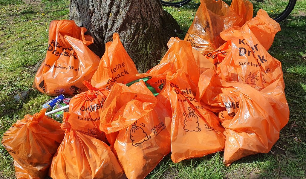 Foto von den organgenen gefüllten Müllsäcken der Stadt Karlsruhe. Es liegen über 10 volle Säcke auf dem Rasen vor einem Baum.