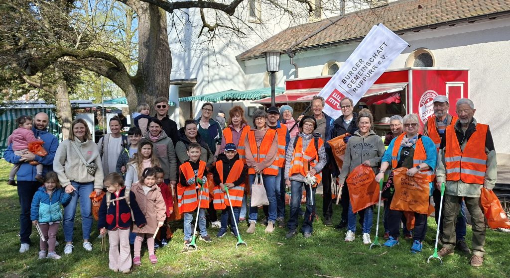 Gruppenfoto von knapp 30 Erwachsenen und Kindern, die an der Sammelaktion teilgenommen haben. Viele davon mit orangenen Sicherheitswesten und Zangen in der Hand zum Müllaufheben. Im Hintergrund die Beachflag der Bürgergemeinschaft Rüppurr.