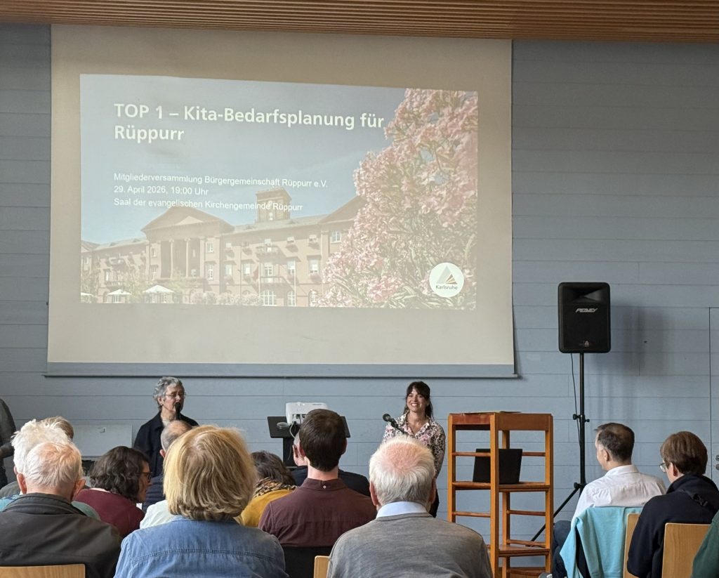 Foto von der Mitgliederversammlung. Vorne sieht man Stuhlreihen mit Zuhörenden. An der Wand hängt eine Leinwand auf der die Präsentation der Stadt für die Kita Bedarfsplanung gezeigt wird. Zwei Mitarbeiterinnen der Stadt Karlsruhe stehen als Referent vor der Leinwand. 
