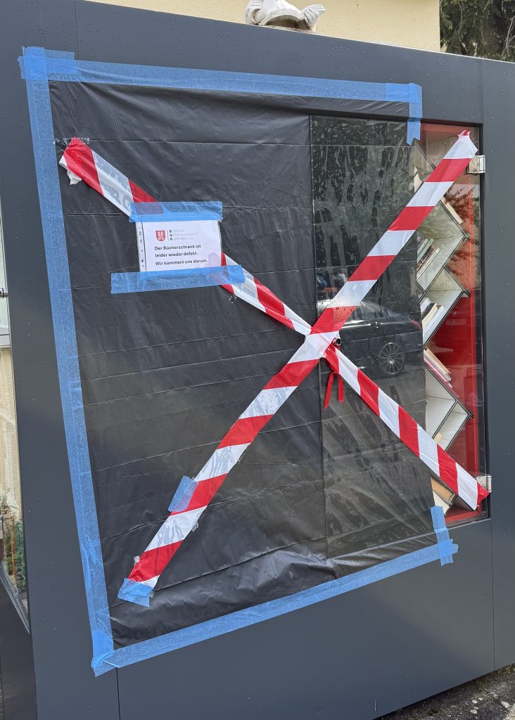 Bild des Bücherschranks auf dem die vorderen zwei Türen mit einer Folie abgeklebt wurden. Darüber ein rote-weißre Flatterband und ein laminierter Hinweis, dass der Bücherschrank erneut defekt ist.