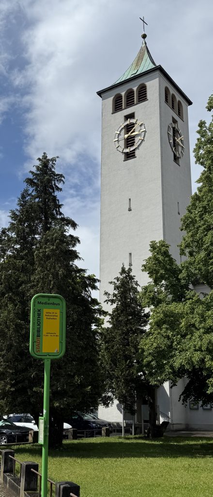 Foto des grünen Schildes für den Medienbus der Stadtbibliothek. Darauf steht, dass der Bus immer mittwochs zwischen 15 und 16:25 Uhr kommt und deshalb hier nicht geparkt werden darf. Im Hintergrund ist der Kirchturm der Christkönig Kirche zu sehen. Dazwischen große grüne Bäume umgeben von Grass.