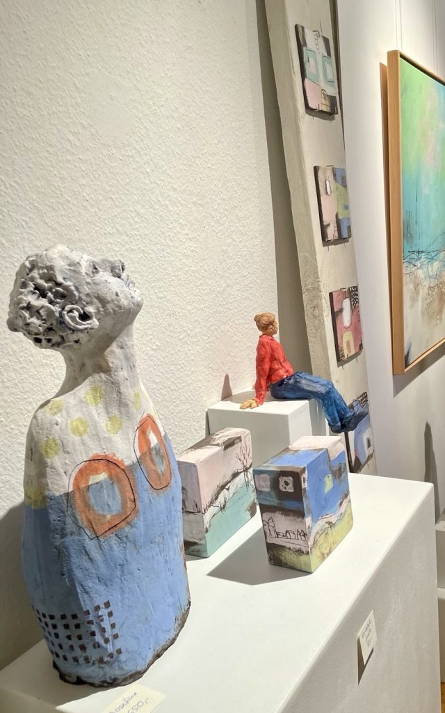 Mehrere Ausstellungsstücke. Ganz links die Figur einer Frau. Daneben 2 bemalte Quader, dahinter eine Figur die auf einem Quadrat sitzt.