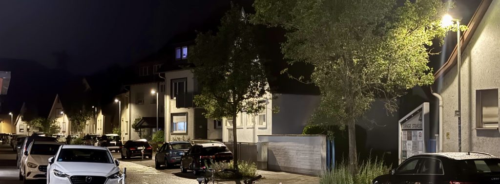 Foto der Lange Straße in Rüppurr. Es ist Nacht. Auf beiden Seiten der Straße parken Autos. Man sieht entlang der rechten Straßenseite die neu aufgestellten Laternen. Die Laternen stehen sehr nah an den Häusern, wodurch diese alle hell erleuchtet sind.