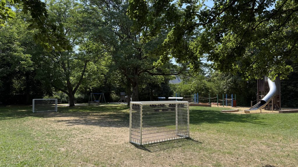 Foto vom Spielplatz. Ansicht mit den neuen Toren im Vordergrund. Im Hintergrund weitere Spiel- und Klettergeräte. Das kurz gemähte Rasenspielfeld ist relativ klein. Vor den Toren ist kein Gras mehr.