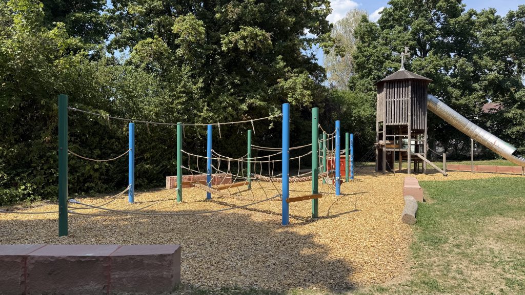 Foto vom Spielplatz. Die neuen Spiel- bzw. Klettergeräte sind zu sehen. Auf dem Boden neu ausgelegte Holzschnitzel damit die Kleinen weich fallen. Zwischen 10 senkrechten blauen und grünen Metallpfosten sind Seile gespannt an denen man sich hangeln kann.