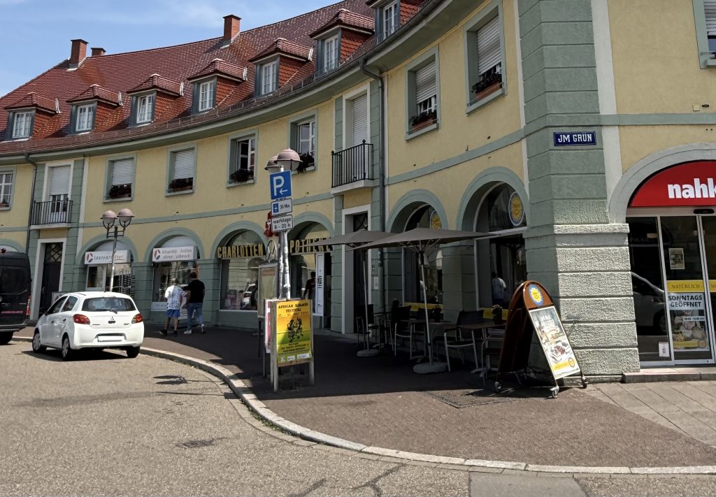 Aufnahme der Ecke Ostendorfplatz und im Grün. Das Gebäude mit dem Nahkauf, der Apotheke und dem Optiker sind zu sehen. Davor ein Parkplatzschild mit der Einschränkung 30 Minuten. Mehrere Autos parken auf der Straße um den Ostendorfplatz.