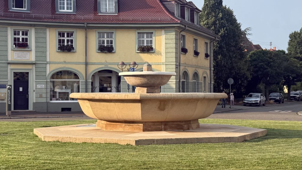 Foto des Brunnen am Ostendorfplatz. Der neu renovierte saubere Brunnen in mitten eines kurz gemähten Rasens. Es ist kein Wasser im Brunnen.