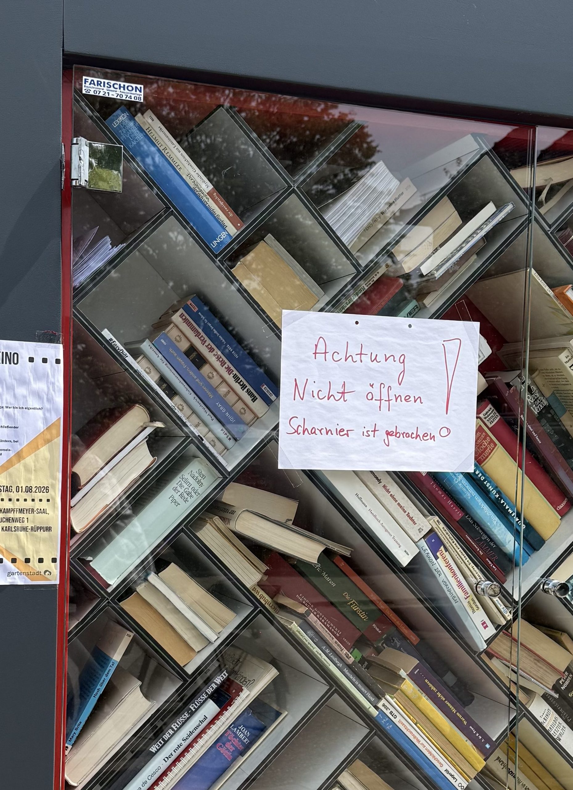 Foto vom Bücherschrank. Ansicht der Glastür. Auf der Tür ist ein Zettel mit der Aufschrift: "Achtung Nicht öffnen. Scharnier ist gebrochen!" Links oben sieht man das defekte Scharnier.