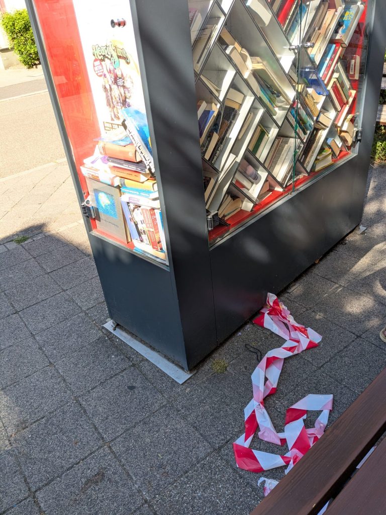 Foto vom Bücherschrank. Auf dem Boden ist das abgerissene rot-weiße Flatterband zu sehen.