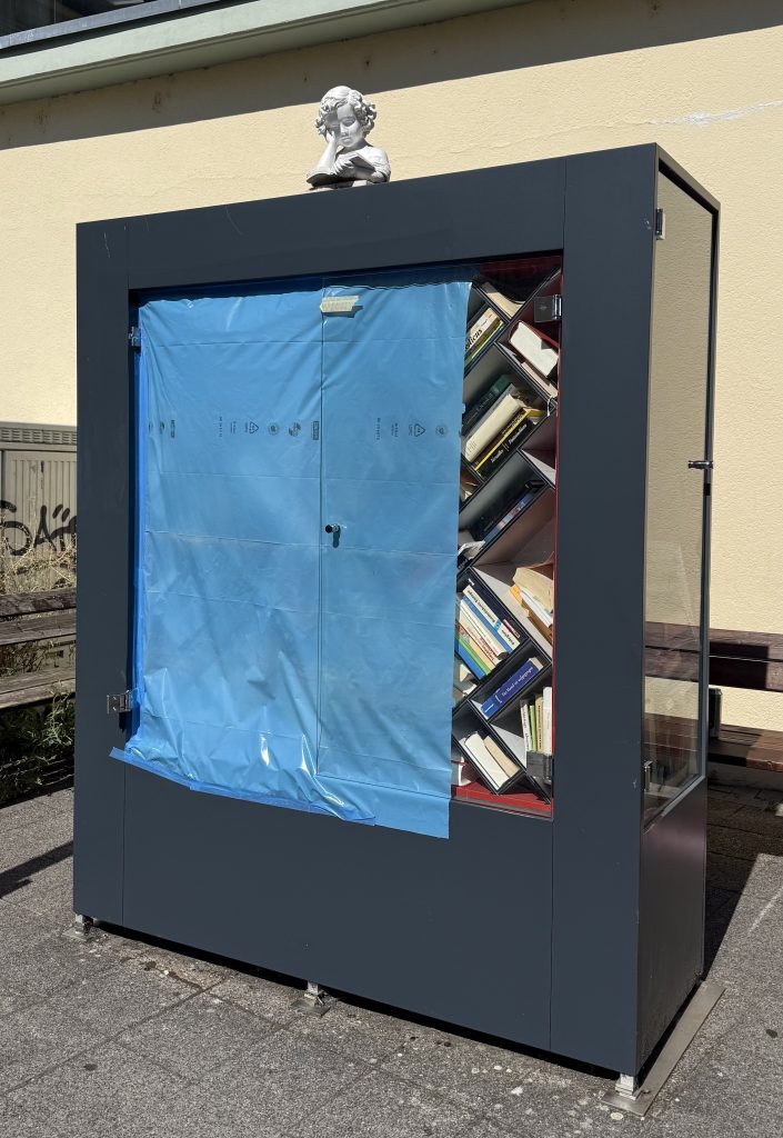 Foto des Bücherschranks. Die linke Glastür wurde entfernt, dafür wurde eine blaue Folie angebracht damit die Bücher nicht nass werden. Die Folie ist fest verklebt, so dass man nicht mehr an die Bücher rankommt.