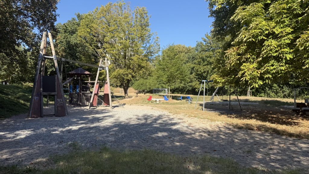 Foto des Spielplatzes. Vorne ist eine größere leere Kiesfläche zu sehen, auf der bisher ein Kletterturm gestanden hat. Links sind noch verbleibende Klettergeräte aus Holz zu sehen. Im Hintergrund sind eine Schaukel und kleine Wippen, dahinter ein Bolzplatz mit Toren.